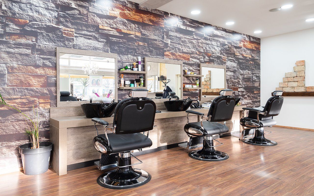 Der Salon in Ismaning: schwarze Friseurstühle vor der Steinwand, Holzboden, Spiegel mit Ablagen.